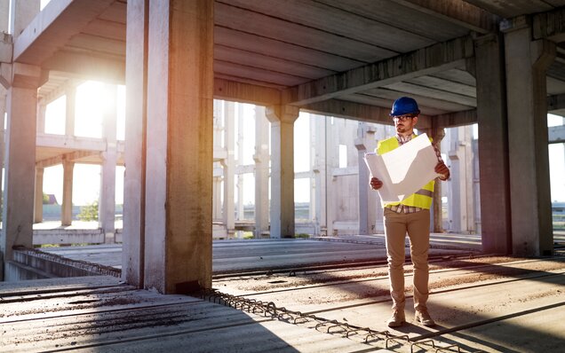 Bauingenieur mit Helm und Sicherheitsweste prüft Baustellenpläne in einem mehrstöckigen Rohbau bei Sonnenuntergang